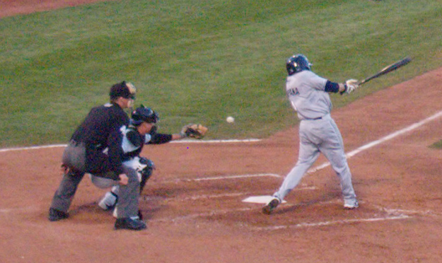 Close up crop of a photo showing a swing and miss, with baseball in view, taken by an Olympus E-P3 in moderately dim light.